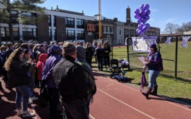 Sarcoidosis Awareness Month | Sarcoidosis News | photo shows Kerry in front of a banner and purple balloons, speaking to a crowd at a high school track