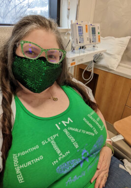 Photo of Kerry at infusion center with a green shirt that uses words like "feeling tired" as letters to spell out the word FINE. She's also wearing glasses with green frames and a sparkly green face mask.
