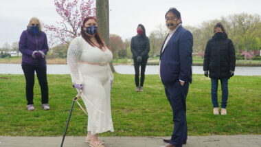 An outdoor wedding photo showing a bride (using a cane), groom, and three attendees, all socially distanced and wearing face masks. The bride and groom appear to be on a sidewalk, with the attendees behind them on green grass. A body of water is behind them.
