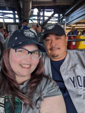 A woman smiles in a dark blue baseball cap, glasses, and a gray T-shirt with a green design. She's beside a man in a dark blue baseball cap and a dark blue T-shirt, over which there's an unbuttoned gray New York Yankees jersey. Metal grids are above them. 