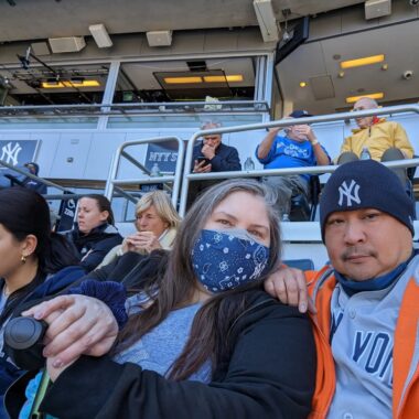 A woman with long dark and gray hair wears a blue face mask with white designs, a dark blue coat, and a light blue shirt. Beside her is a man wearing a New York Yankees dark blue stocking cap, a gray Yankees jersey, and an orange coat. Both are seated in a stadium with other people and a New York Yankees symbol in the background.