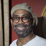 A headshot of Charlton Harris shows him smiling and wearing a white shirt, knitted hat, and glasses.