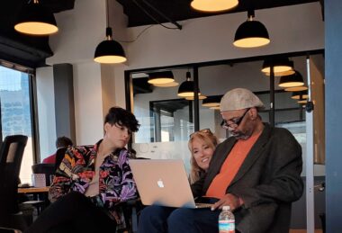 Charlton Harris sits on a chair with a computer on his lap. He's wearing a gray suit jacket, orange shirt, and what appears to be an Ivy cap. Two co-workers, both women, are seated to his right and looking at his computer screen. They're in an office with several lights hanging overhead.