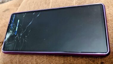 A close-up photo of a cracked cellphone in a purple case lying on a wooden table. The screen is completely black.