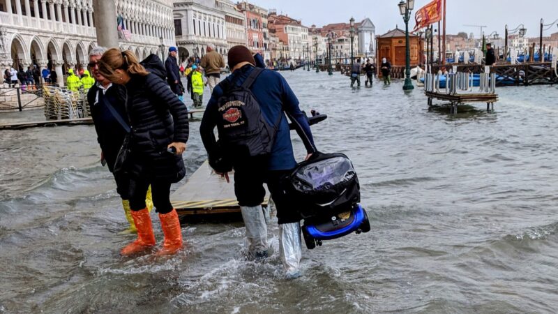 Photo shows a people walking through a flooded street in Venice, Italy. At center, a man is carrying a folded mobility scooter in the air.