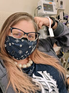Photo of Kerry wearing Yankees glasses, mask, and shirt, large pearl necklace, and hoodie with zipper opening in the sleeve, through which her IV connects to her port.