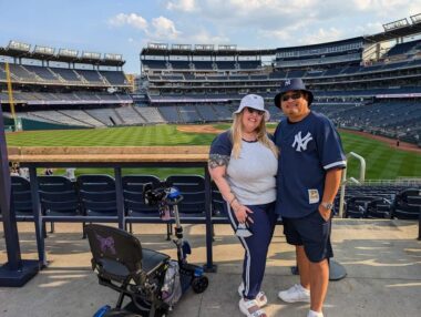 Photo of Kerry and her husband dressed in New York Yankees gear, standing next to her travel scooter in a baseball stadium.