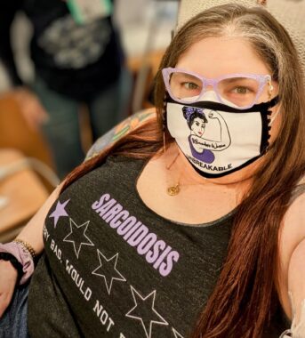 Photo of Kerry at her infusion center, wearing a "Sarcoidosis Warrior: Unbreakable" mask and a tank top with one-star review that reads, "Sarcoidosis: Very bad, would not recommend"